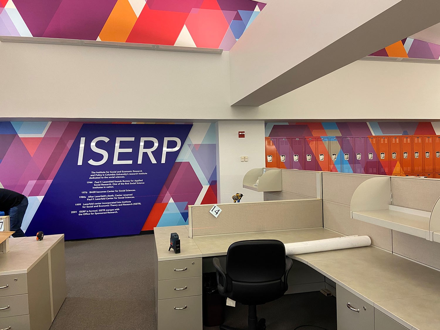 "After" photo of the ISERP space. A colorful, zig-zag pattern of custom designs covers the formerly white wall, with the ISERP logo and mission statement on the centerpiece of the design.